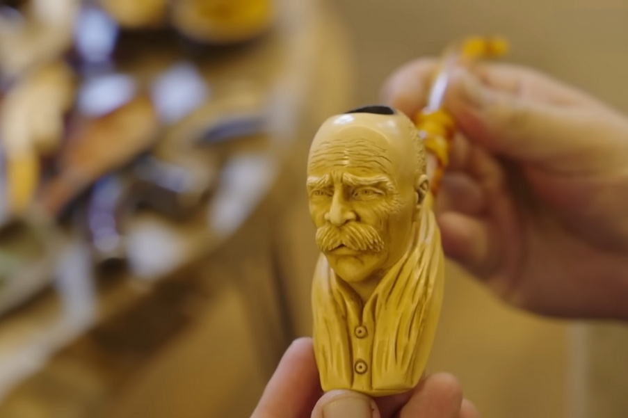 Hand-carved meerschaum pipe featuring a detailed portrait of an elderly man with a mustache and scarf, held in hand with blurred pipes in the background.
